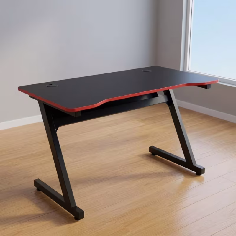 Ergonomic PC Gaming Desk featuring a matte black desktop with a curved front edge, a protective red trim, dual cable grommets, and a black steel Z-shaped leg frame.